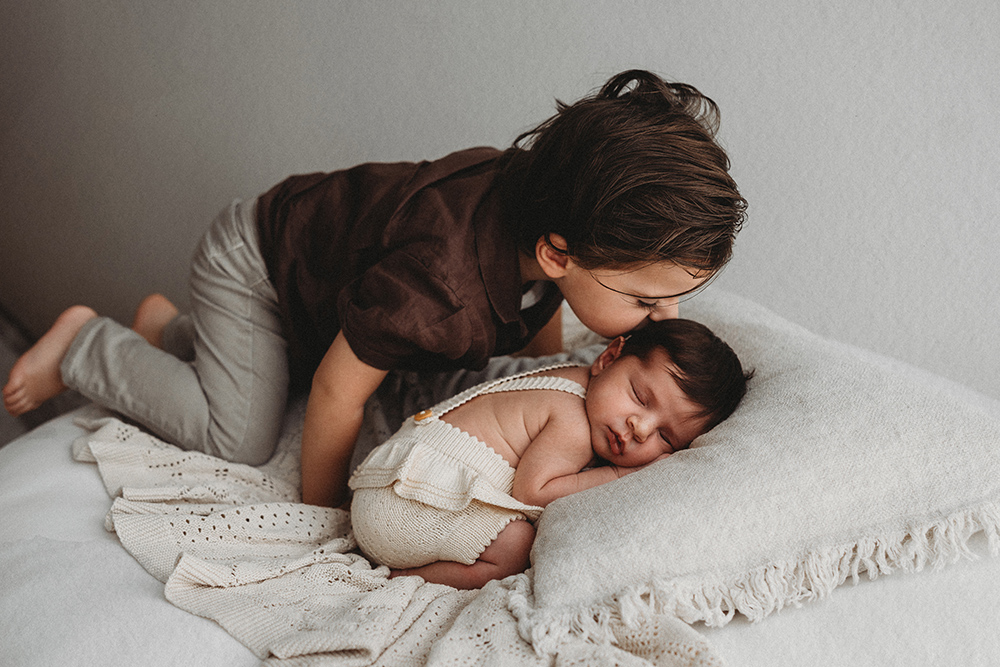 Broertje geeft kus aan slapende newborn tijdens newbornshoot in daglichtstudio.