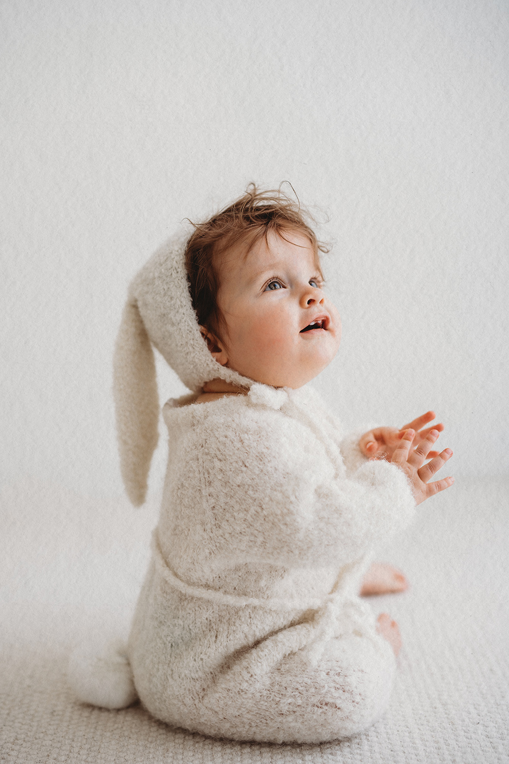 Baby in witte outfit tijdens een sittershoot in daglichtstudio, zittend en omhoog kijkend.